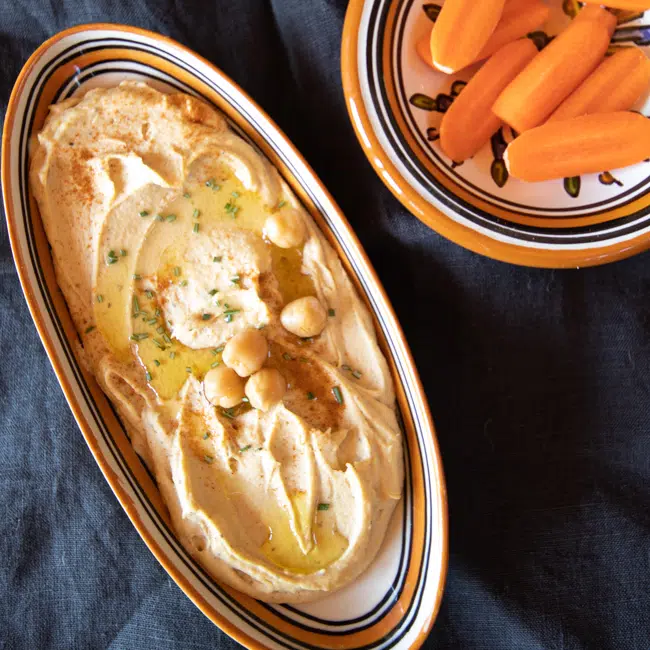 Creamy hummus dip in a serving dish, topped with a few chickpeas and chives, served with carrots.