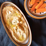 Small dish of creamy smooth hummus with a sprinkling of paprika, and a few chickpeas. Some raw carrots are on the side to dip into the hummus.