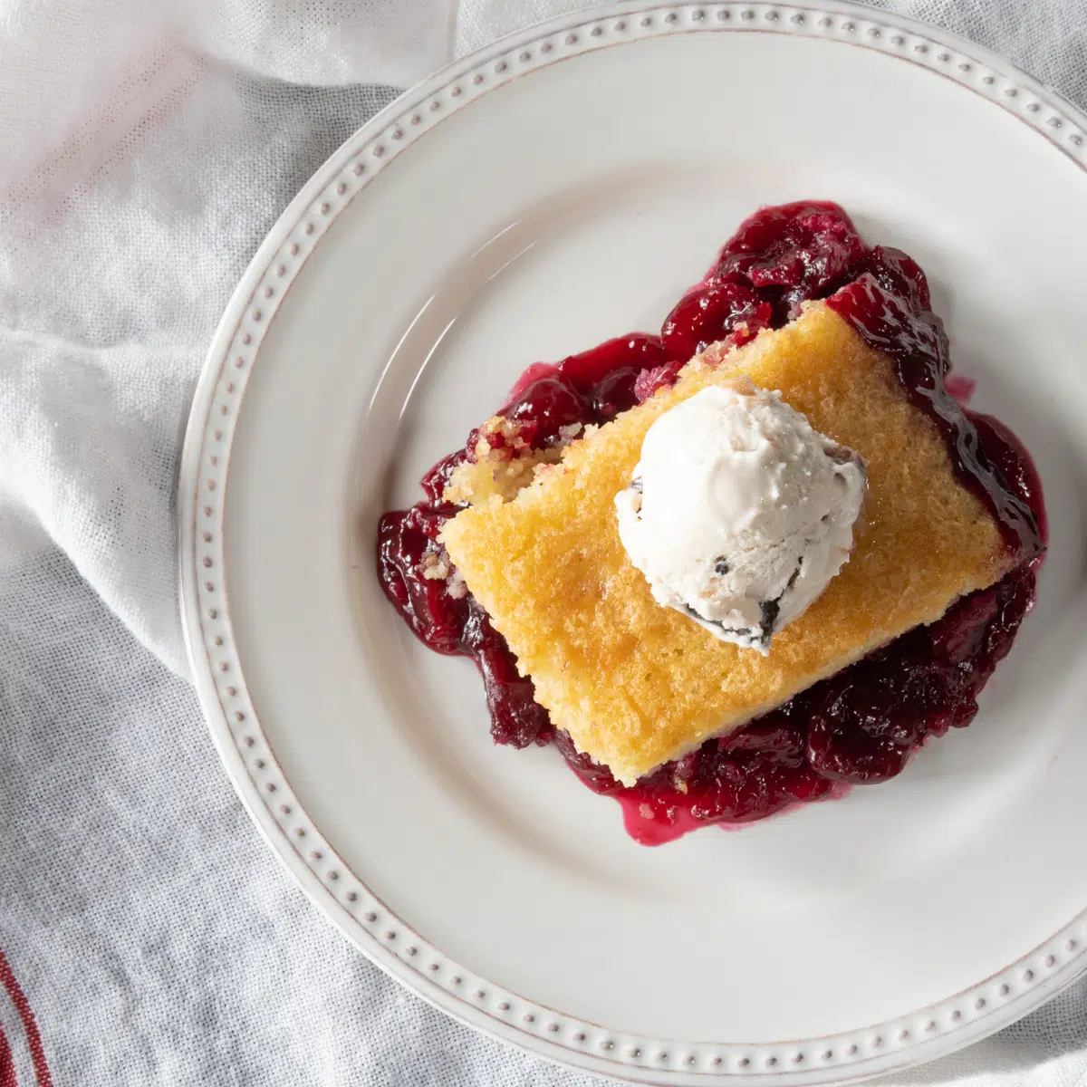Cherry cobbler with a cake-like topping on a plate, topped with a scoop of ice cream.