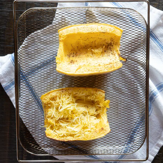 Spaghetti squash halves in the basket of an air fryer, with one of the halves all fluffed up with a fork showing the spaghetti strands.