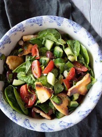 Bowl of fattoush salad with baby spinach greens, tomatoes, kalamata olives, cucumbers, crispy chickpeas and crisp pita bread pieces.