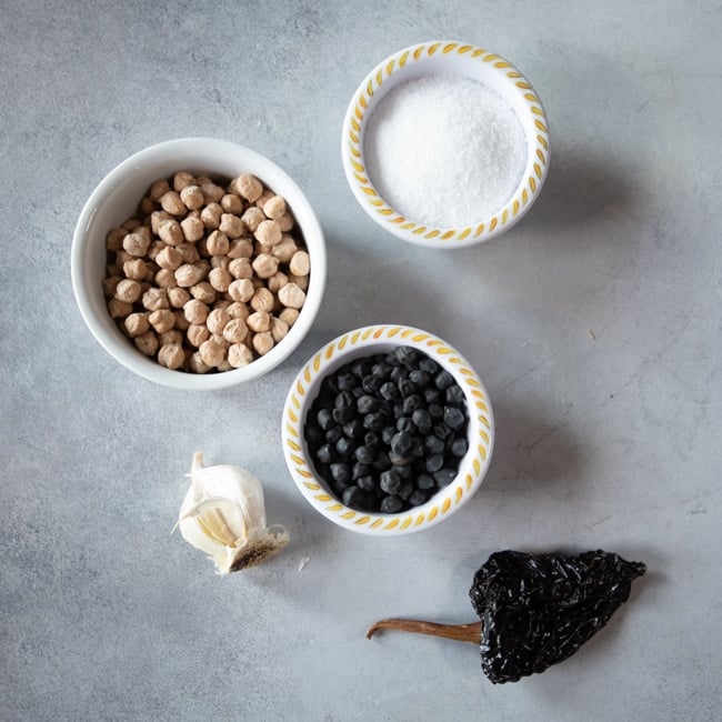 Small bowls of tan chickpeas, black chickpeas and kosher salt, with garlic and a dried pepper on the side.