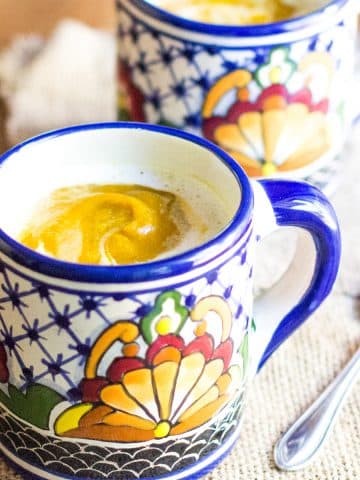 A cup of pumpkin soup topped with foamed Half & Half, sitting on a placemat with a spoon.