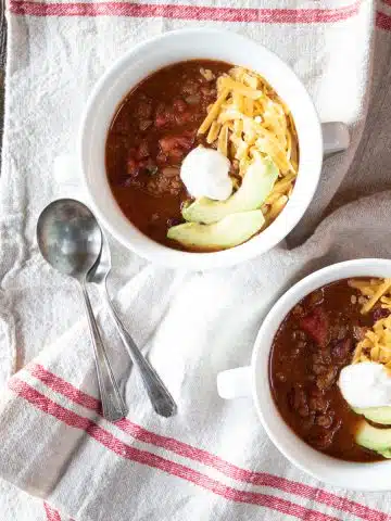 Two bowls of vegan chili con carne topped with avocado slices, vegan sour cream and vegan cheddar cheese.