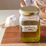 A labeled jar of garlic oil, with a head of garlic and breads in the background.
