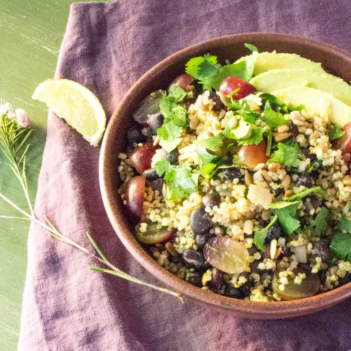 A bowl of quinoa, black beans, grapes, avocado and cilantro, on a tea towel with a slice of lime.