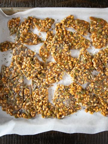 Pieces of pine nut and pumpkin seed brittle broken up on a baking sheet.