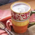 A cup of hot cocoa topped with coconut whipped cream, and 2 spoons.