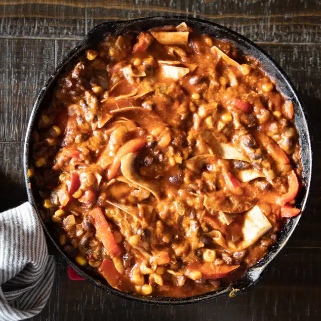 A cast iron skillet filled with skillet enchiladas with red and green peppers, black beans, corn, enchilada sauce and tortilla shards.