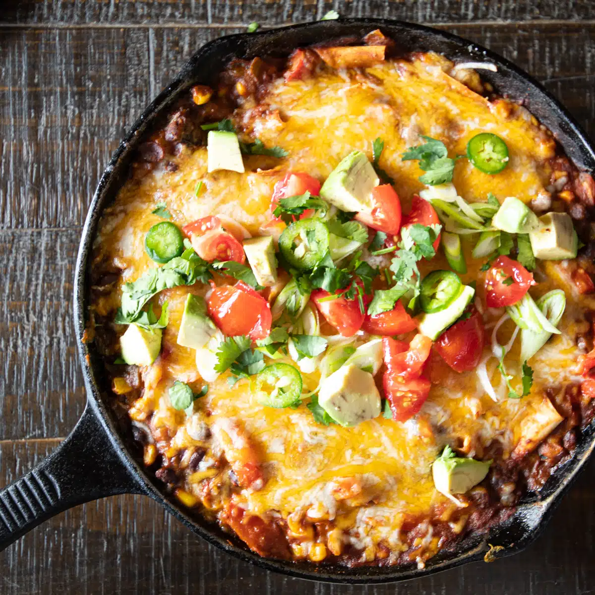 A cast iron skillet of skillet enchiladas topped with cheese, avocado, tomatoes, scallions, cilantro and jalapeño slices.