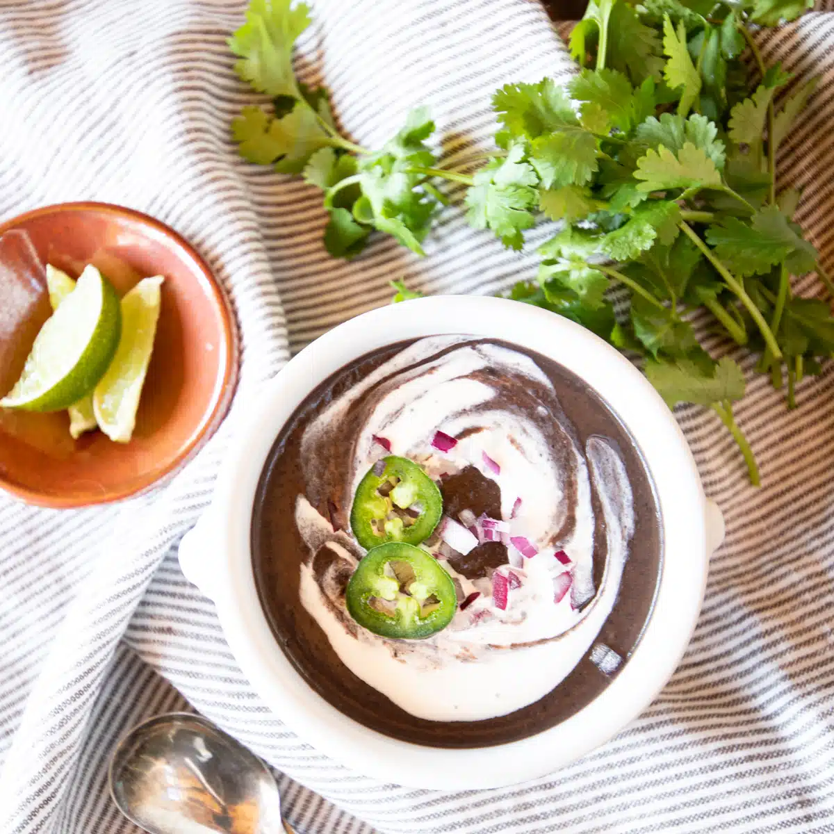 Bowl of black bean soup with sour cream, red onion and jalapeno slices, with lime wedges and cilantro off to the side.