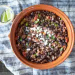 Mexican black beans topped with cilantro and grated cheese in a dish, with lime slices on the side.