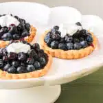 Three blueberry tartlets glazed with a blueberry glaze, and topped with whipped cream, on a dessert server.
