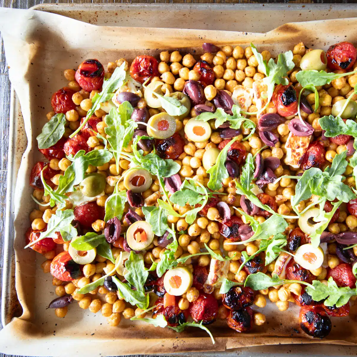 Warm roasted chickpea feta salad with olives, fire roasted tomatoes and arugula on a baking sheet ready to be served.