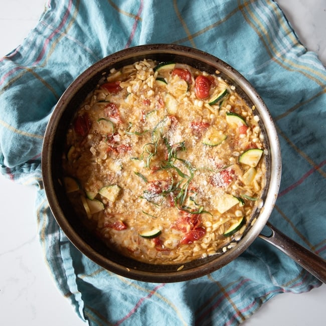 One-pot pasta dish of tomatoes, zucchini, corn, pesto, and mozzarella mixed into orzo in a skillet topped with fresh basil and Parmesan cheese.