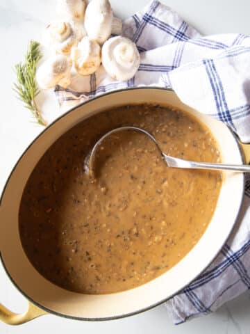A big pot of mushroom gravy with a few mushrooms and a sprig of rosemary beside it.
