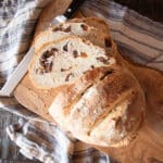 A baked round of fig bread on a cutting board with a bread knife. The bread is sliced into a few slices to show the figs in the bread.